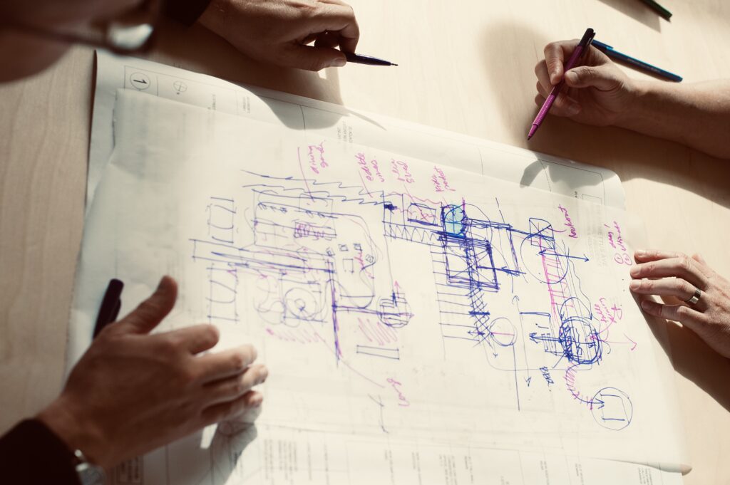 Three people sketching and discussing architectural plans with pens on a large sheet of paper.
