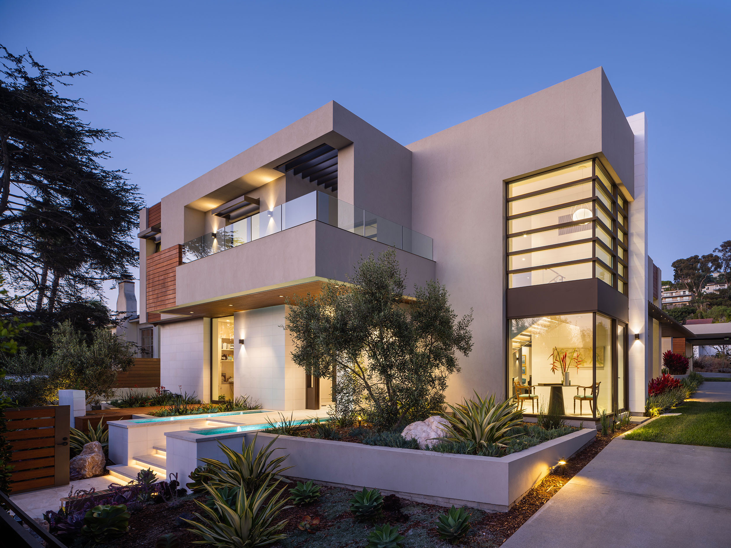 Modern two-story house with large windows, balcony, garden, and illuminated exterior at dusk.