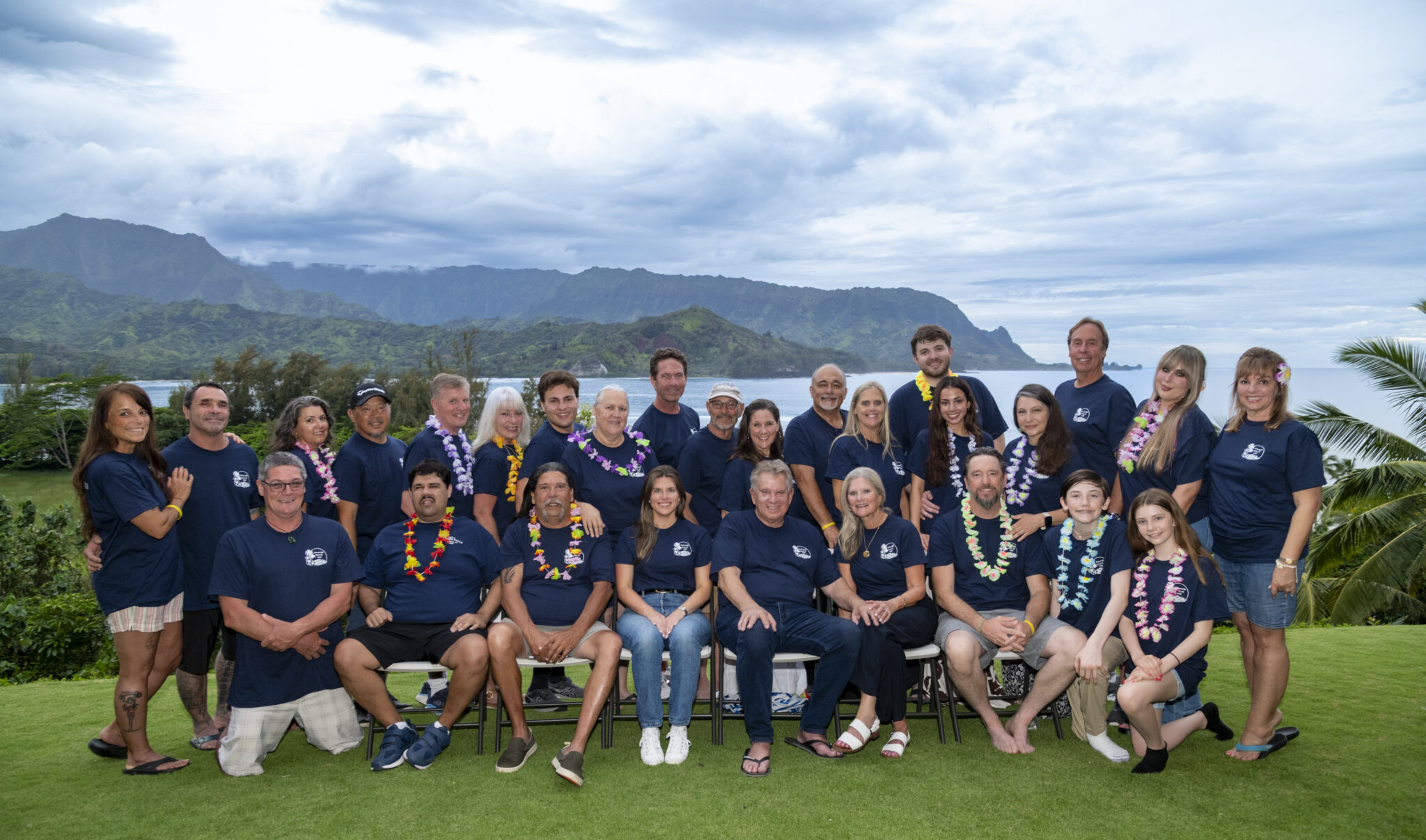 A group of people in matching blue shirts pose outdoors with mountains and greenery in the background.